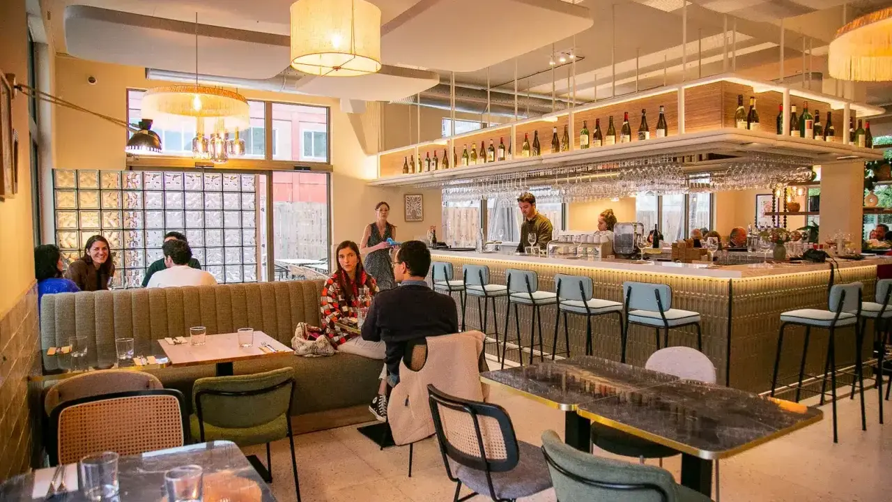 Ambiance chaleureuse d'un restaurant, avec des clients attablés et un bar bien garni. L'esprit du terroir se retrouve dans la sélection des vins et la convivialité.