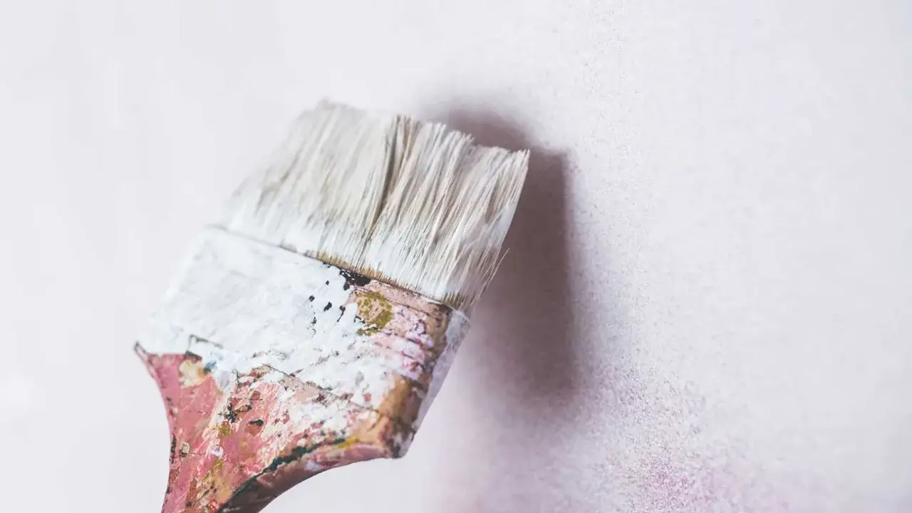 Pincel desgastado con restos de pintura blanca a punto de tocar una pared de cal con textura sutil.