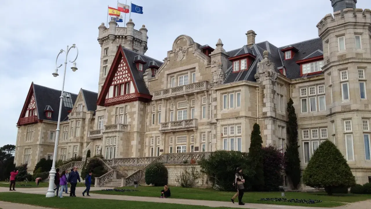 Palacio de la Magdalena en Santander, con banderas ondeando en la torre. Turistas pasean por los jardines de esta joya arquitectónica.