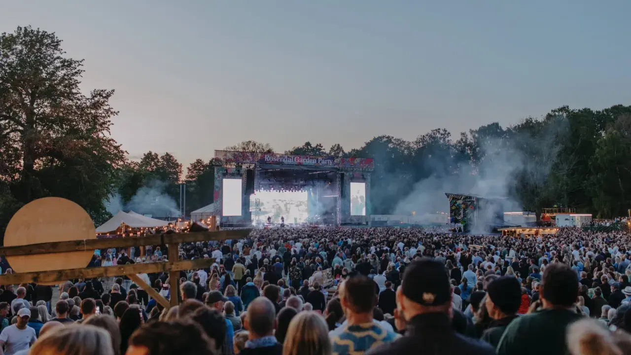 Stor publik vid Rosendal Garden Party. En stor scen med artister och ljus. Rök och träd i bakgrunden.