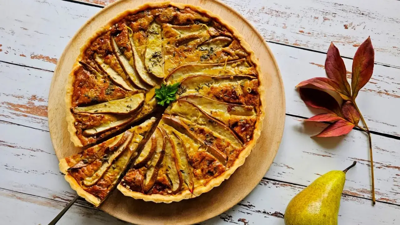 Apetyczna tarta z gruszkami, udekorowana natką pietruszki i skórką cytrynową, prezentuje się na drewnianej desce. Obok leży świeża gruszka i jesienne liście.