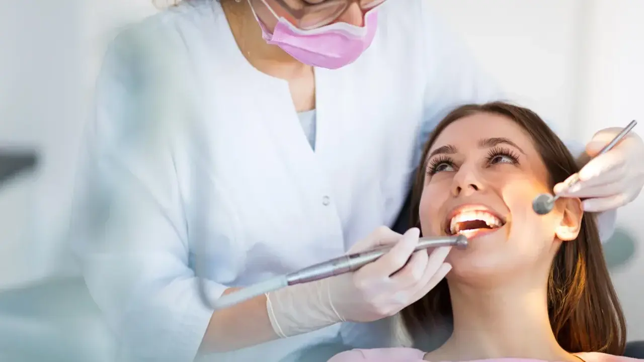 Dentystka w masce i rękawiczkach bada zęby pacjentki za pomocą lusterka i wiertła.
