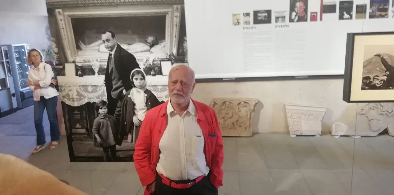 Un uomo anziano con giacca rossa, davanti a una foto di Ferdinando Scianna in Sicilia con bambini.