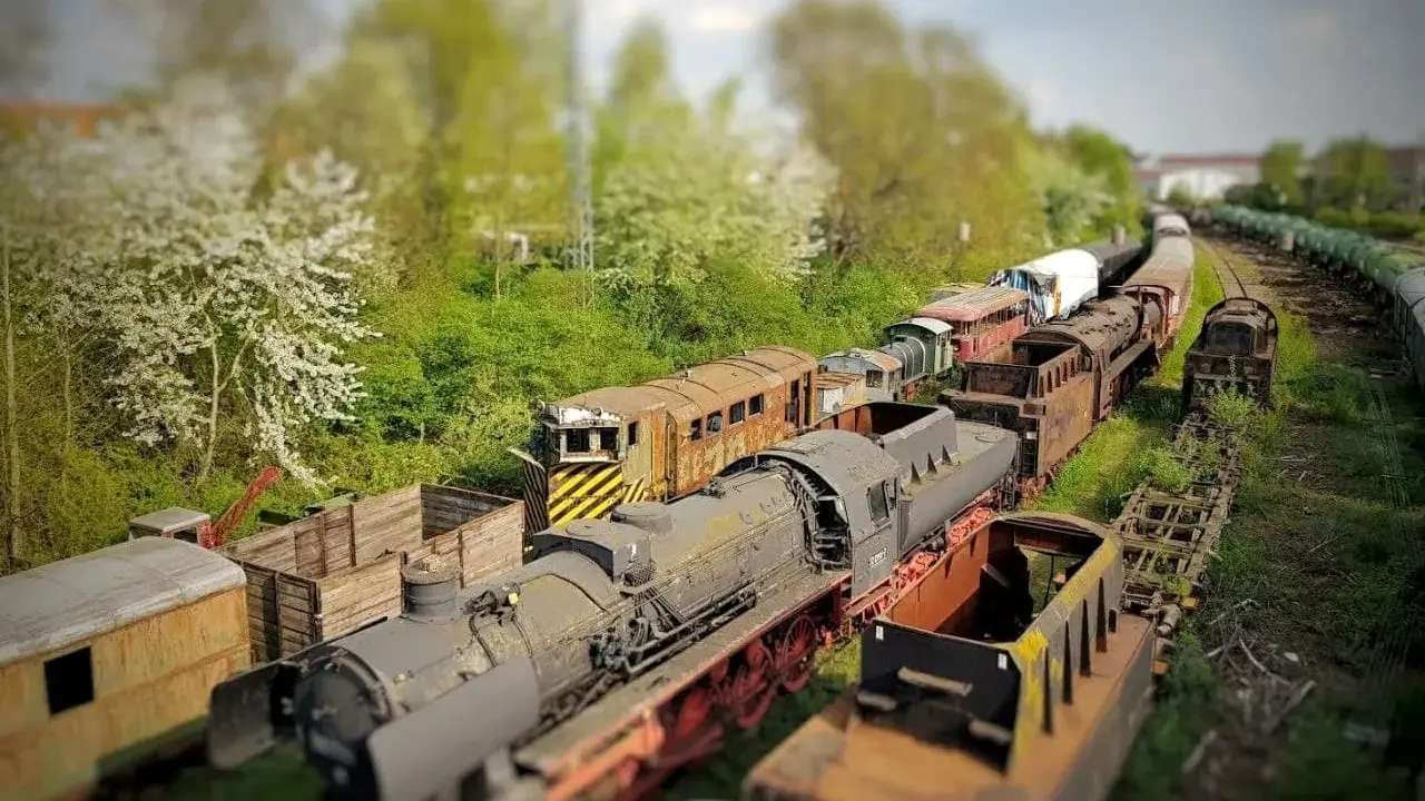 Un cementerio de trenes con efecto tilt shift, donde locomotoras y vagones oxidados parecen miniaturas bajo un cielo nublado.