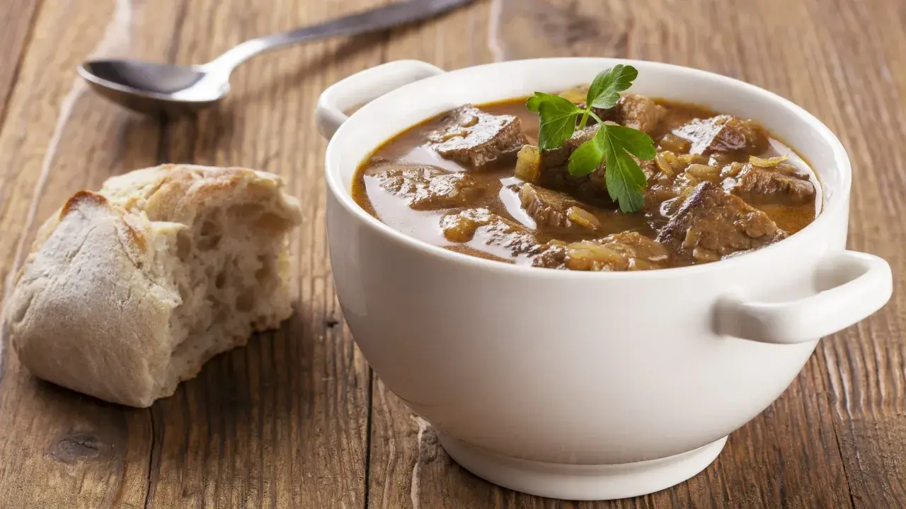Aromaticzny, tradycyjny gulasz wołowy w białej miseczce, z gałązką pietruszki, obok kromka chleba i łyżka.