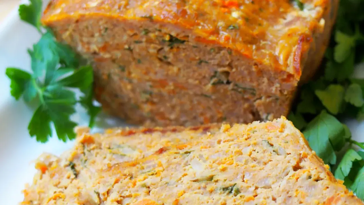 Pyszna pieczeń z mięsa mielonego, pokrojona w plastry, z widocznymi kawałkami marchewki i ziół, udekorowana natką pietruszki.