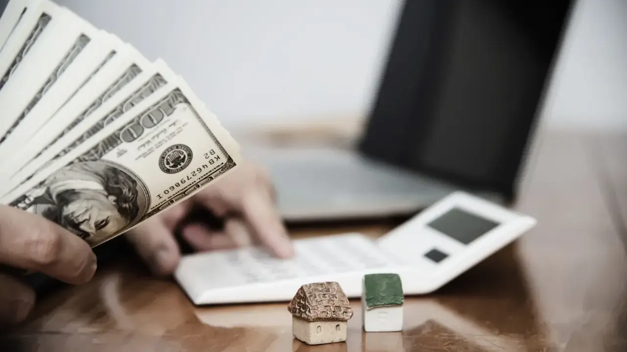 Ręka trzyma banknoty 100 USD, obok kalkulator, laptop i miniaturowe domki. Zastanawiasz się, ile wynosi zasiłek socjalny na mieszkanie?