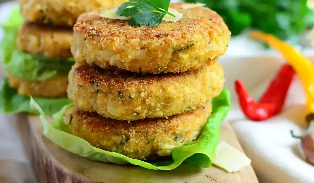 Złociste kotlety z kalafiora na 3 sposoby, podane na liściach sałaty, z parmezanem i natką pietruszki. Obok czerwone i żółte papryczki chili.