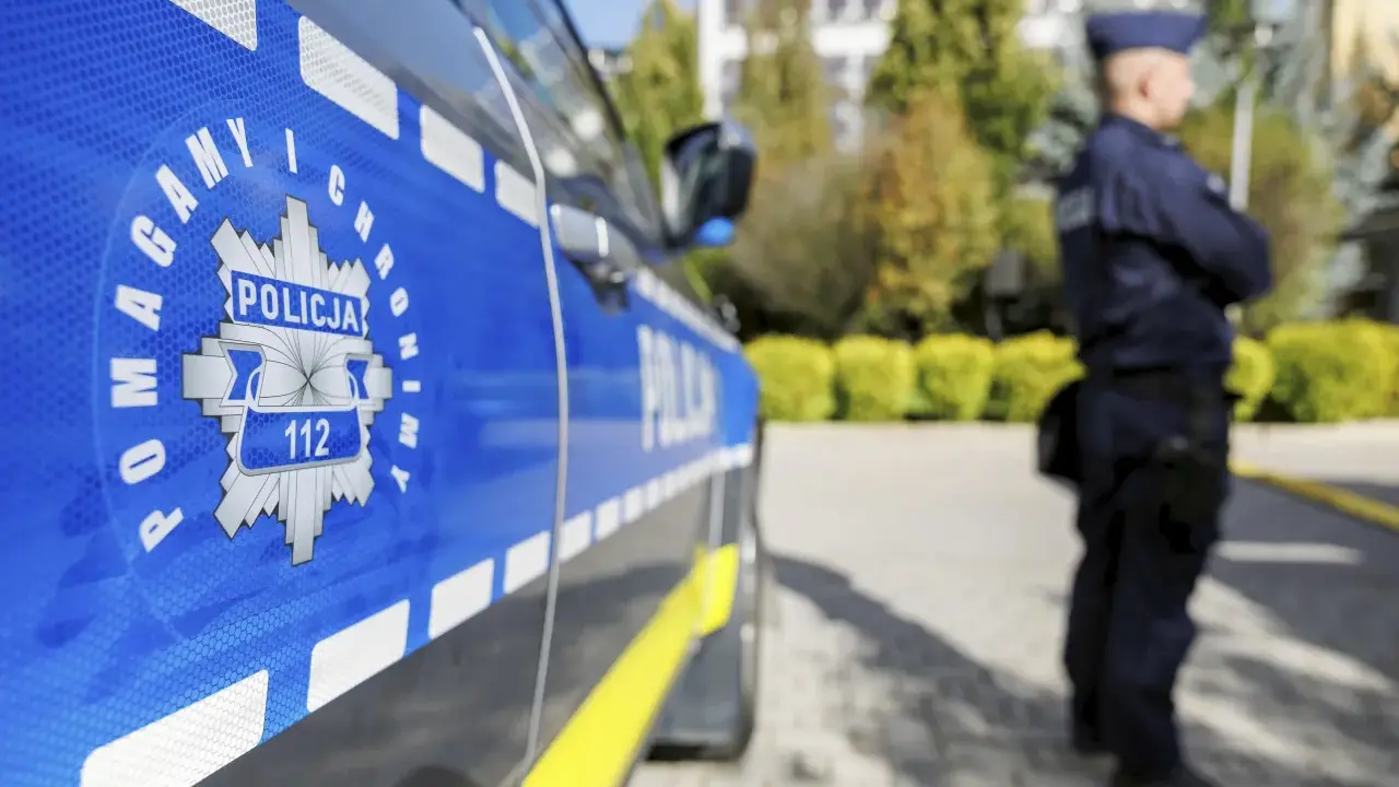 Policjant w mundurze stoi obok radiowozu z widocznym znakiem Policji i numerem 112.