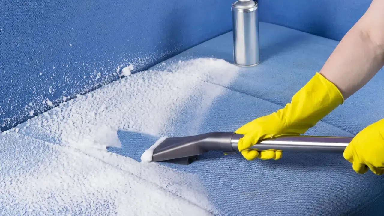 Mani con guanti gialli eseguono il lavaggio divano a secco con schiuma e aspiratore su tessuto blu.