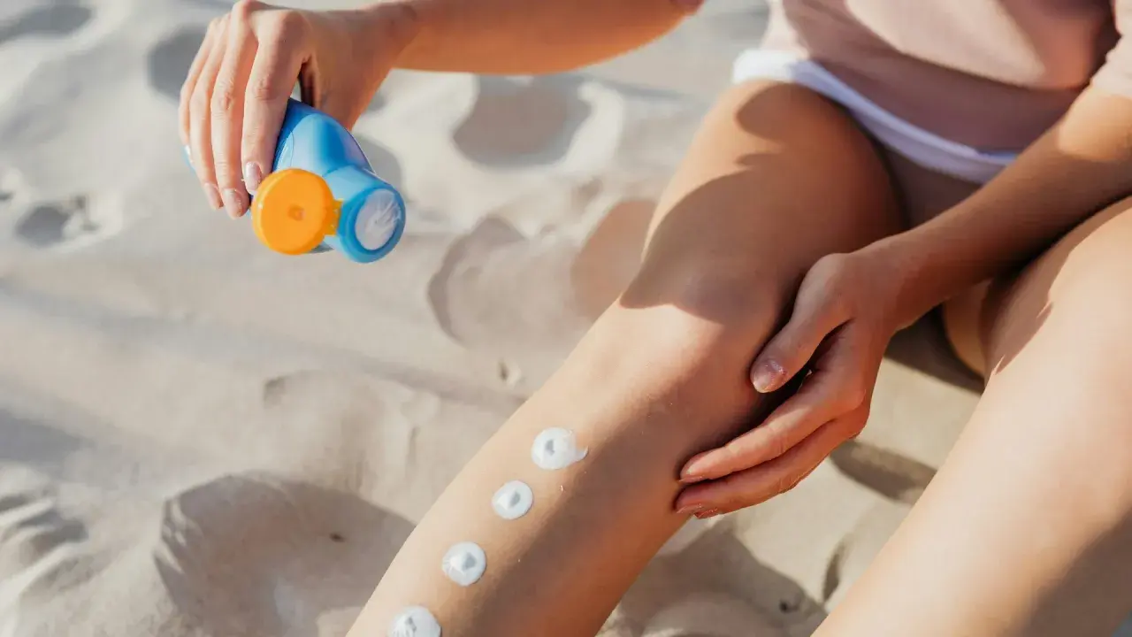 Main sur le sable, une main applique des points de crème solaire sur une jambe. Attention, cette crème solaire perimee pourrait ne plus protéger efficacement.