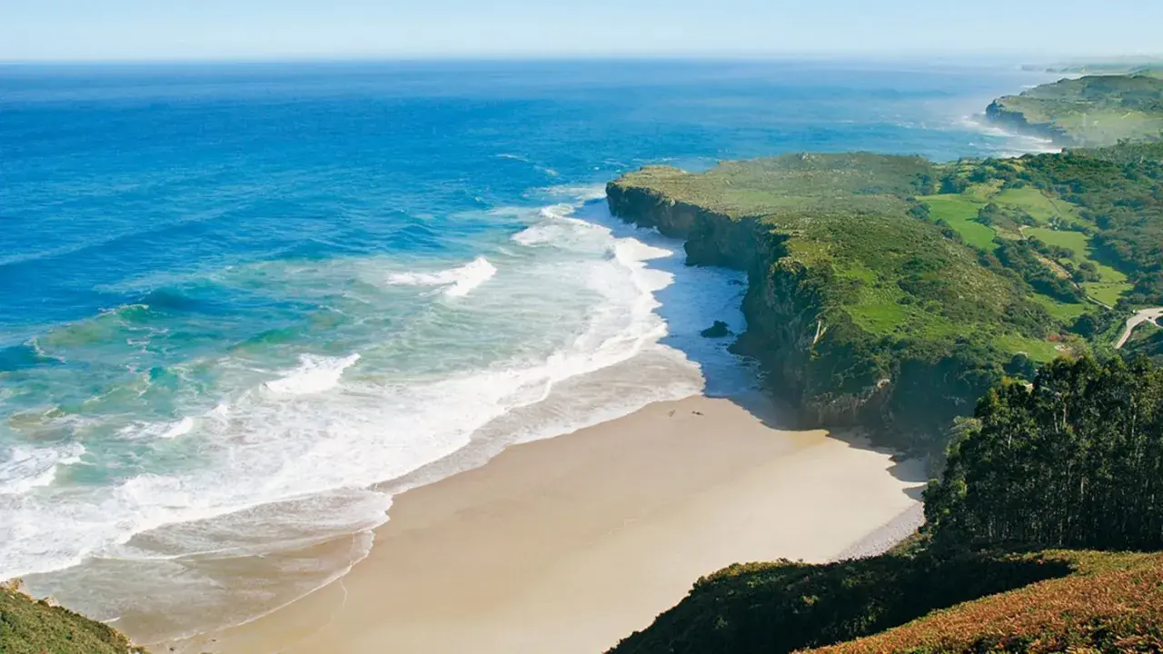 Atemberaubende Küstenlandschaft am Camino del Norte. Grüne Hügel treffen auf das azurblaue Meer mit sanften Wellen, die auf einen einsamen Sandstrand treffen.