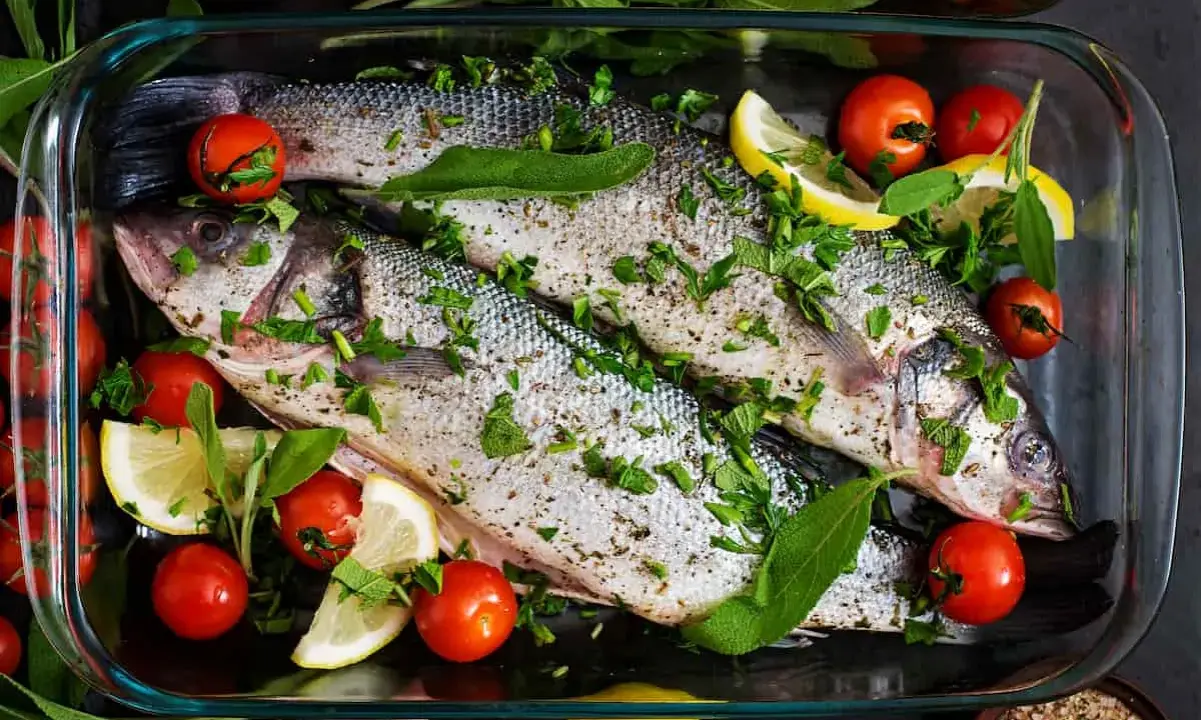 Dos lubinas enteras preparadas para ser horneadas, con tomates cherry, rodajas de limón y hierbas frescas en una bandeja de vidrio.