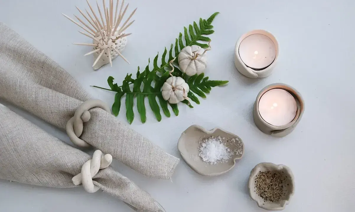 Två ljusstakar av lufttorkande lera med värmeljus, salt- och pepparskålar, servettringar och en dekoration av torkade pumpor på en ormbunksgren.