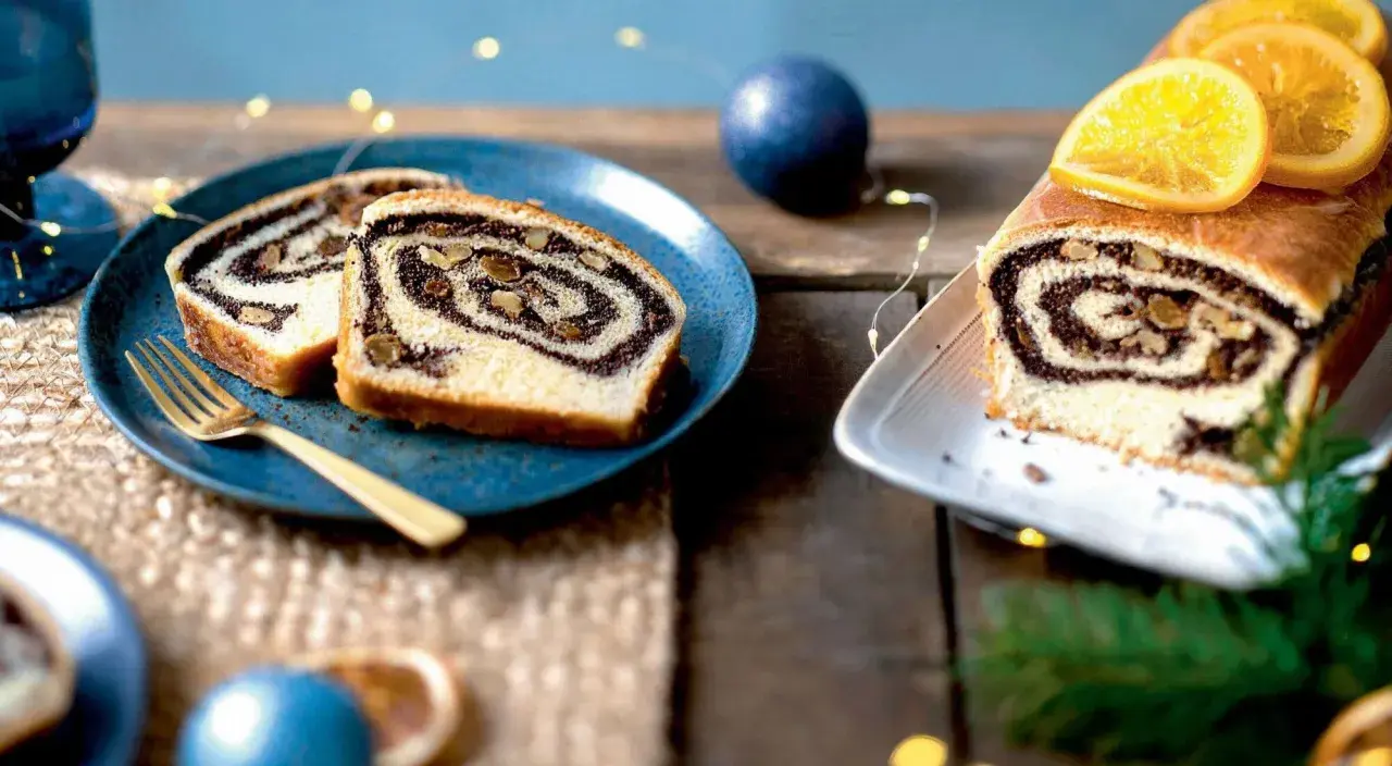 Tranches de roulé au pavot, garni de noix, sur une assiette bleue avec une fourchette dorée. Un autre roulé décoré d'orange est à droite.