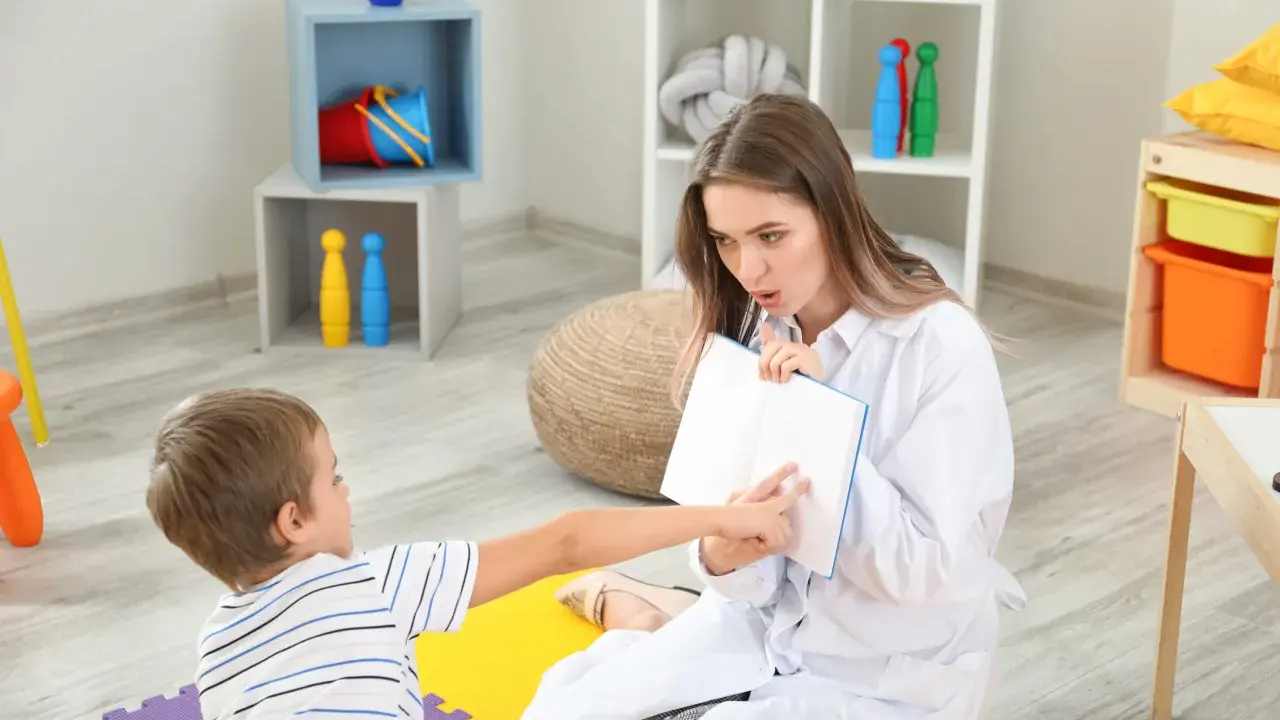 Terapeuta pracuje z dzieckiem nad poprawą niewyraźnej mowy, wskazując palcem na strony otwartej książki.