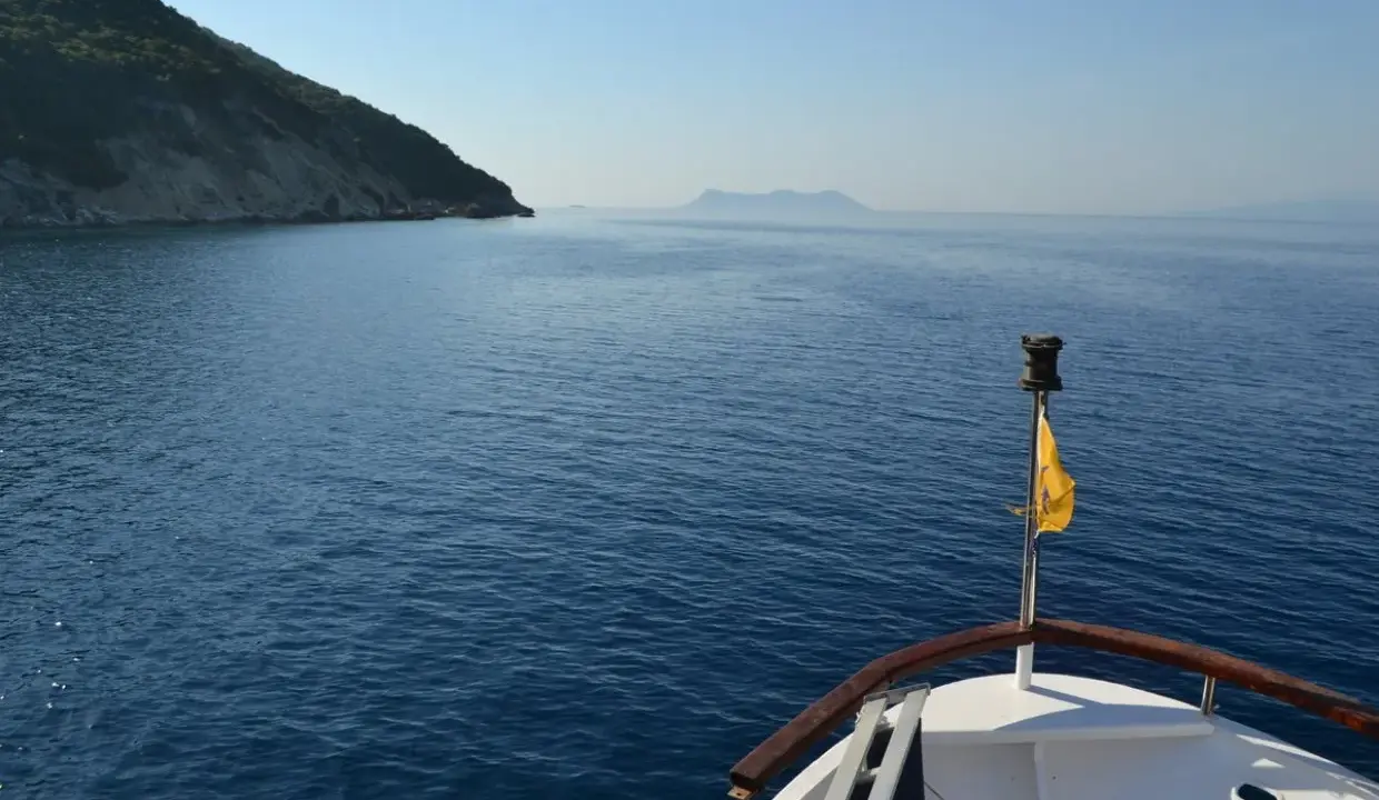 La prua di una barca naviga verso un'isola lontana, un viaggio che evoca la poesia di Itaca.