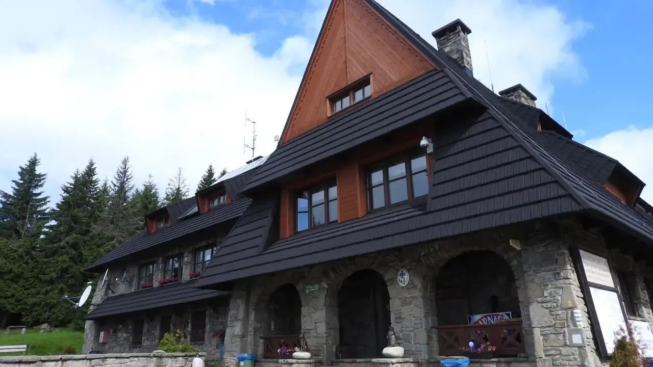 Kamienny schron turystyczny pod gorącymi szczytami, z drewnianą przybudówką i czarnym dachem, otoczony lasem świerkowym.