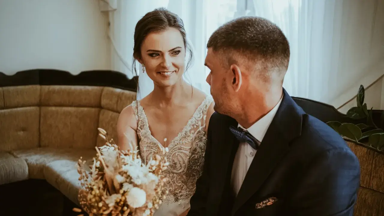 Uśmiechnięta panna młoda w sukni z koronkami i pan młody w granatowym garniturze. W tle kanapa i zasłony. Krótka ceremonia, bo ile trwa ślub cywilny?