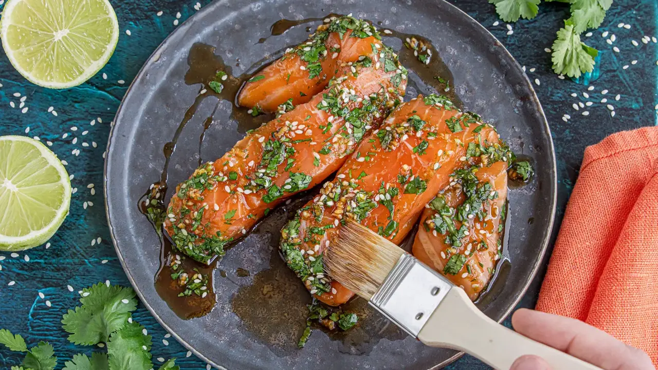 Lachsfilets werden mit einer asiatischen Marinade aus Sojasauce, Ingwer, Knoblauch, Kräutern und Sesam bestrichen. Dazu Limettenspalten und Koriander.