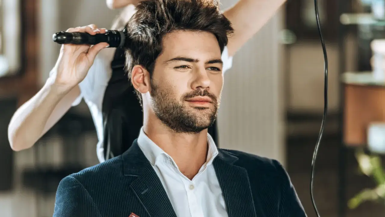 Mężczyzna w eleganckim garniturze czeka na stylizację fryzury męskiej. Fryzjerka suszy jego włosy suszarką.
