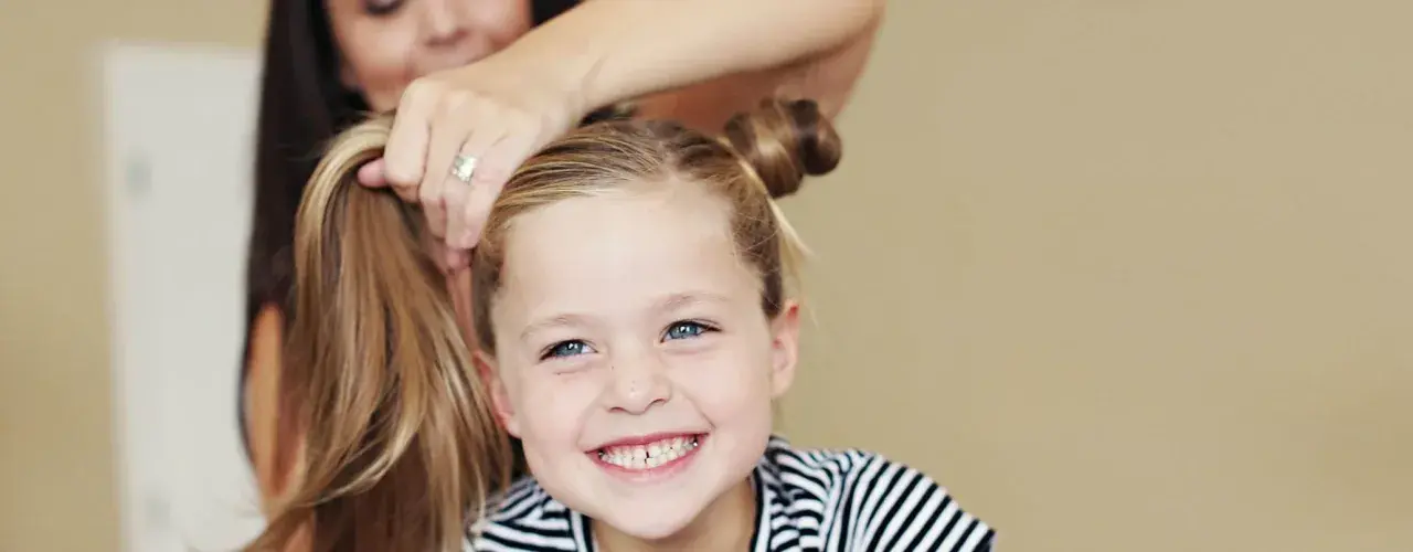 Ein Mädchen mit blauen Augen und Sommersprossen lächelt, während ihre Mutter ihr die Haare zu niedlichen Dutts frisiert. Tolle Ideen für kinder frisuren!