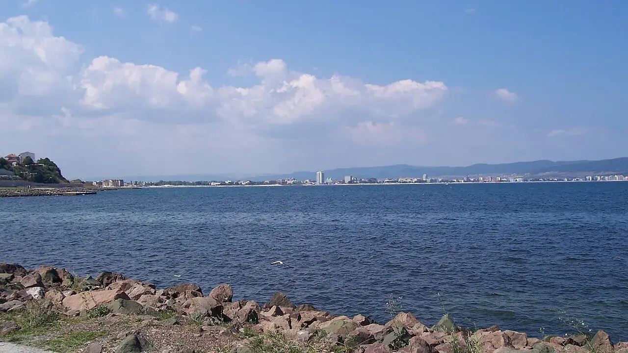 Słoneczny brzeg jak pić. Widok na morze i miasto w oddali, z kamienistym wybrzeżem na pierwszym planie.