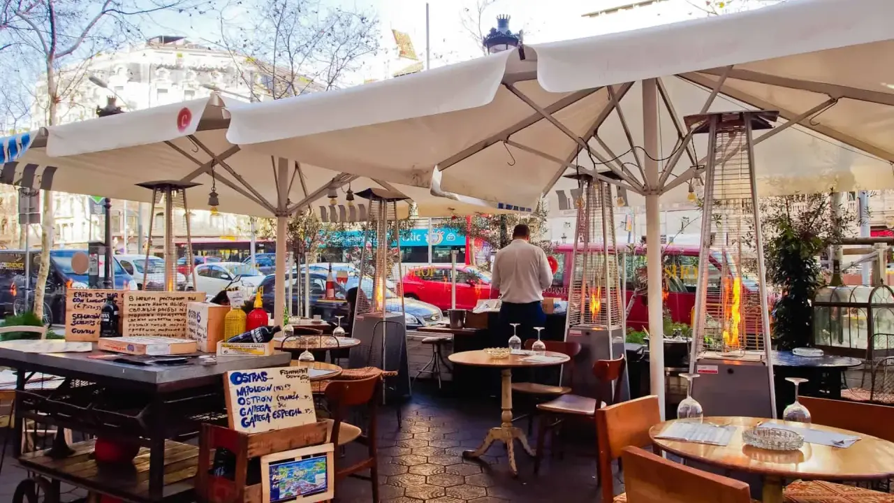 Terrasse d'un restaurant à Barcelone, avec des tables, des parasols, des chauffages d'extérieur et des panneaux indiquant des plats typiques espagnols.