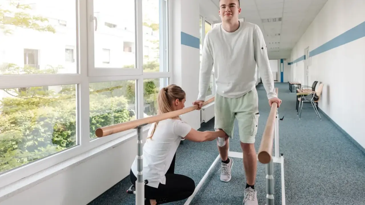 Młody mężczyzna ćwiczy chód przy poręczy, a fizjoterapeutka pomaga mu w rehabilitacji.