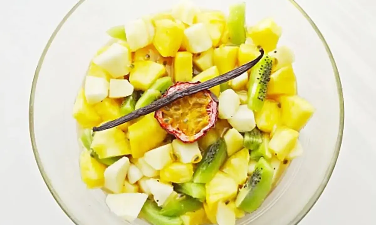 Salade de fruits tropicaux coupés en dés dans un bol en verre, garnie de fruits de la passion et d'une gousse de vanille.