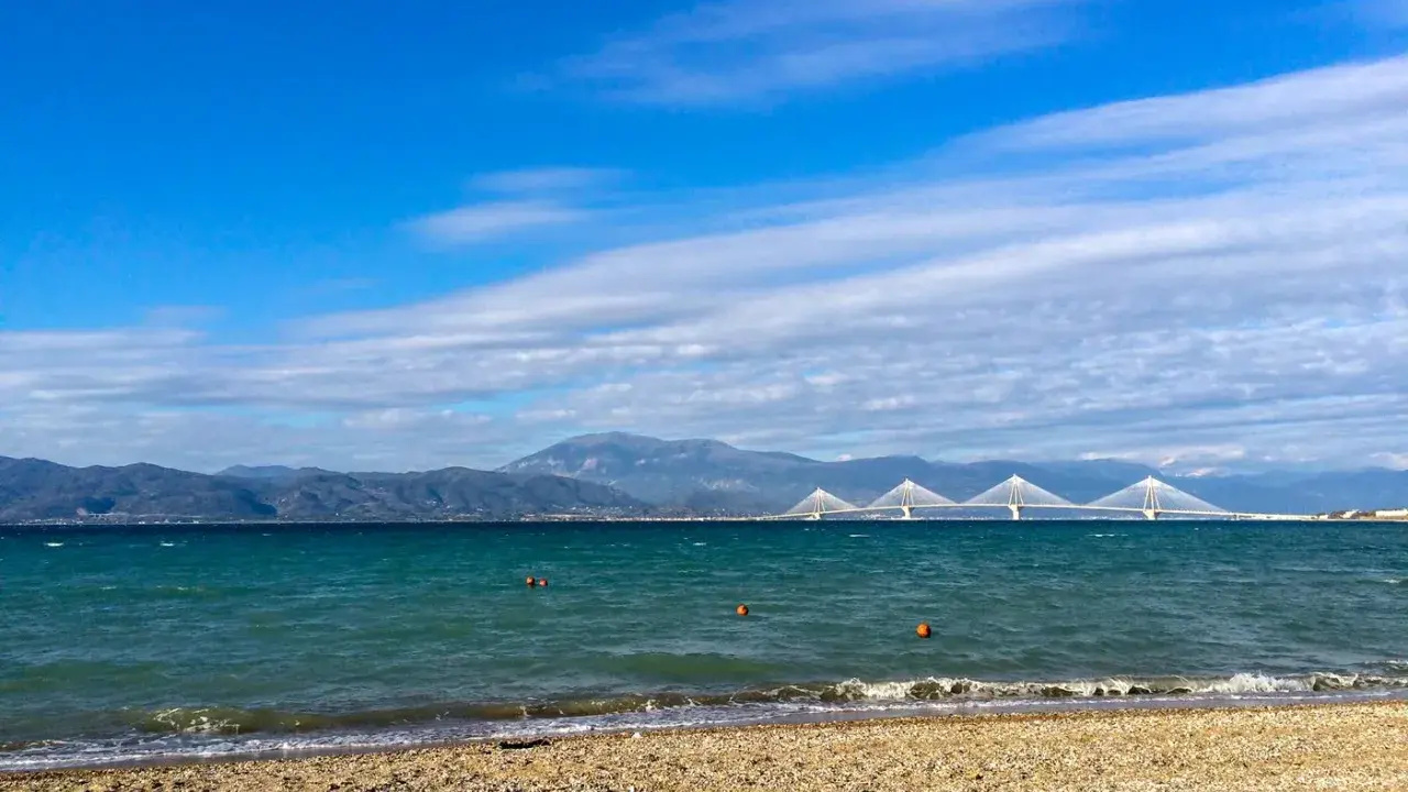 Spokojne morze i plaża w Grecji w styczniu. W oddali widać most Rio-Antirio i góry.