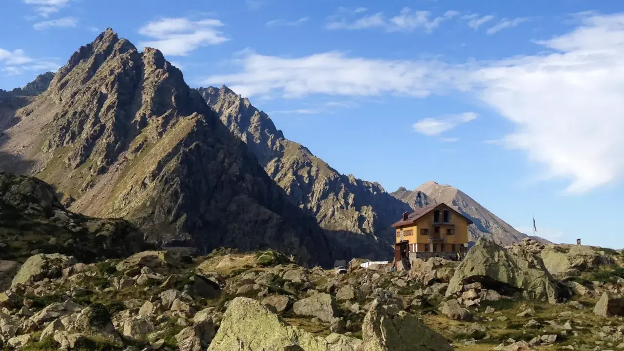 Il rifugio Genova, incastonato tra imponenti montagne rocciose e un cielo azzurro punteggiato di nuvole, offre un riparo solitario e suggestivo.