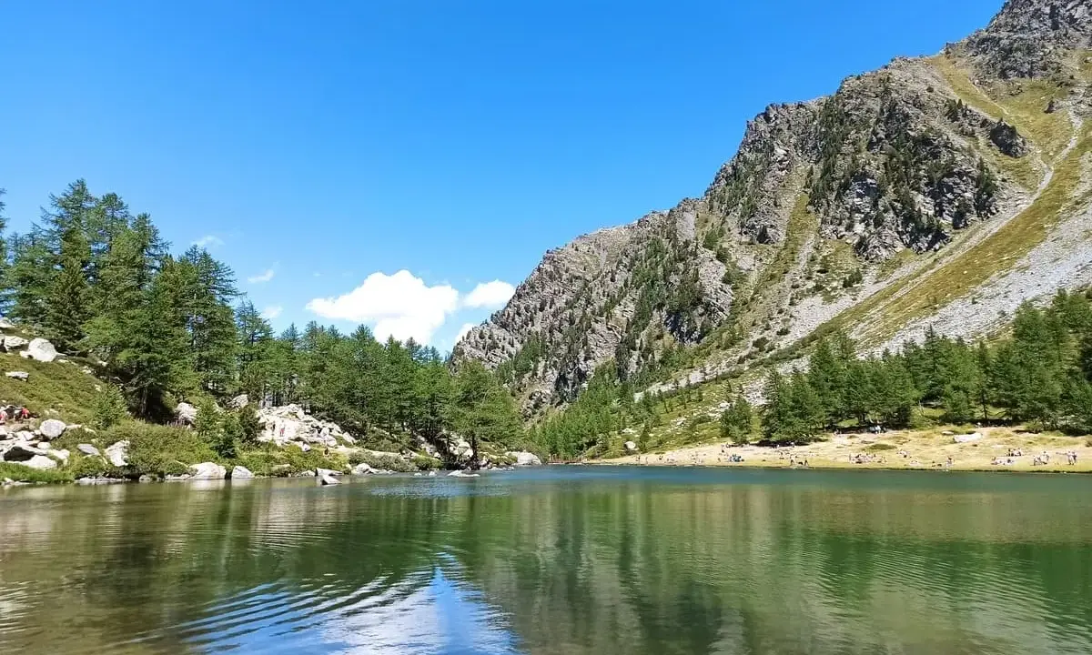 Il **lago d'Arpy** riflette il cielo azzurro e le montagne rocciose. Sulle rive, pini e persone godono del sole estivo.