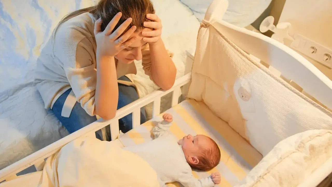 Madre agotada, con las manos en la cabeza, mira a su bebe que se desvela por la noche en su cuna.