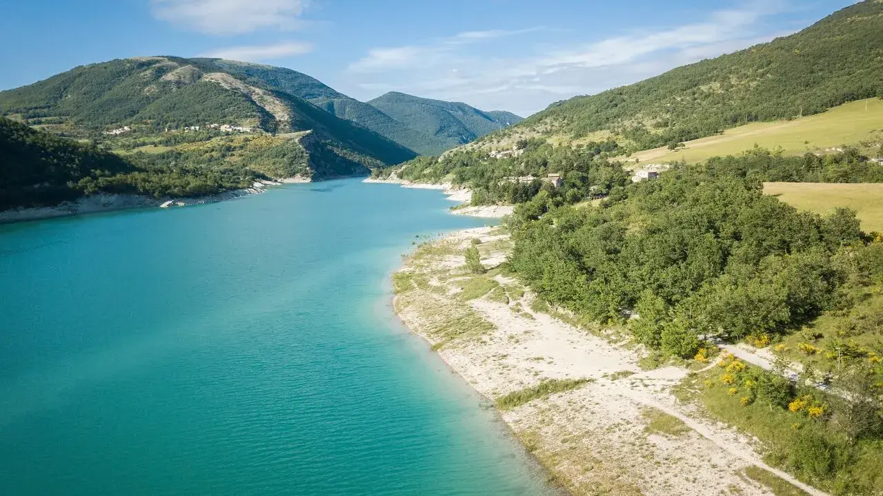 Splendido **lago di Fiastra** con acque turchesi, circondato da colline verdi e case sparse. Un paesaggio sereno e incantevole.