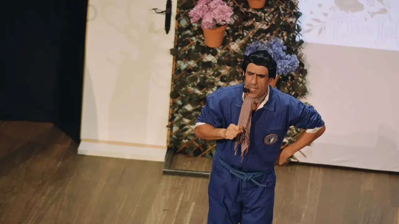 Actor con mono azul y pañuelo a cuadros, actuando en un teatro para niños. Fondo con flores y enredaderas.