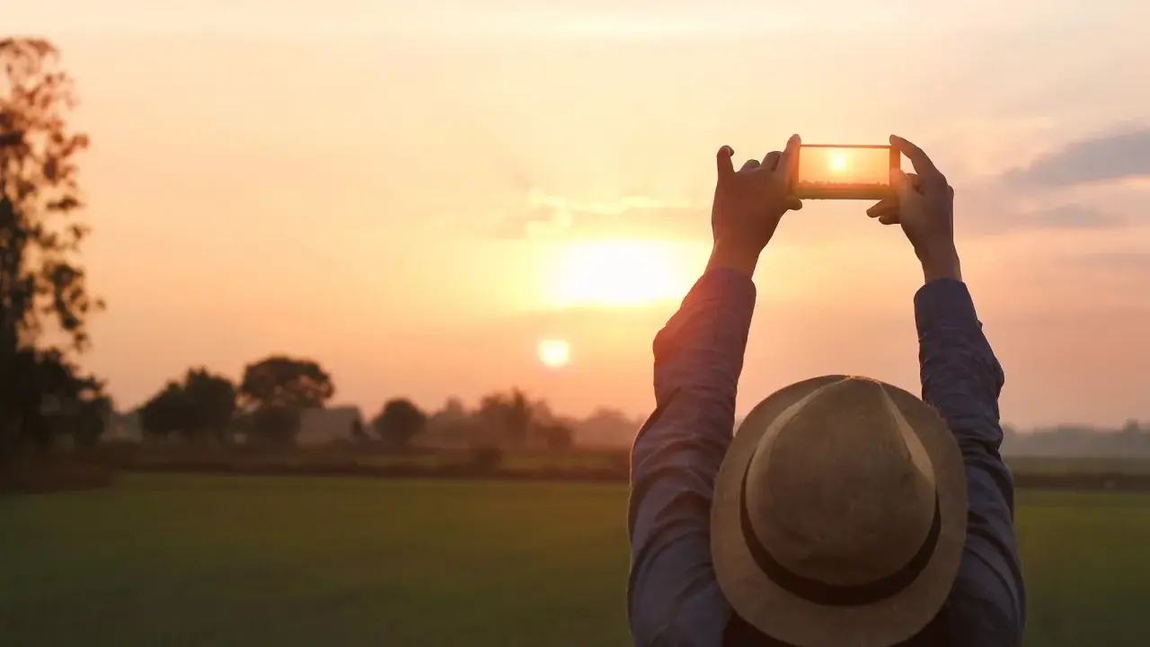 Osoba w kapeluszu fotografuje zachód słońca telefonem z aparatem szerokokątnym.