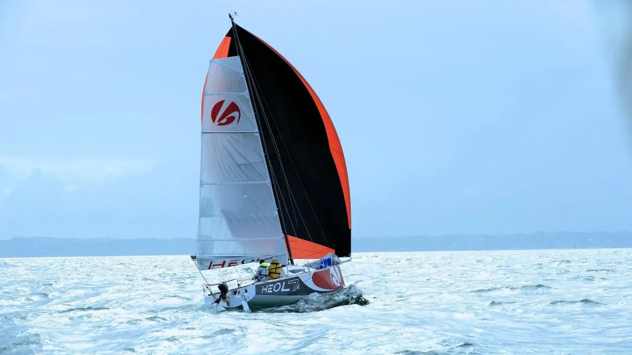 Voilier Heol 7.4 fendant les vagues, ses voiles bicolores gonflées par le vent. Deux marins à bord.