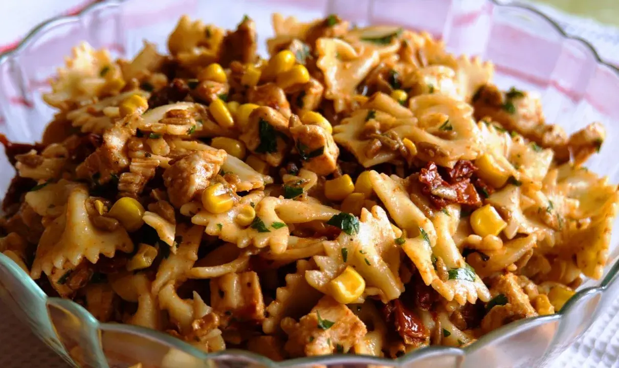 Pyszna sałatka z kurczakiem i makaronem farfalle, kukurydzą i suszonymi pomidorami, posypana natką pietruszki.