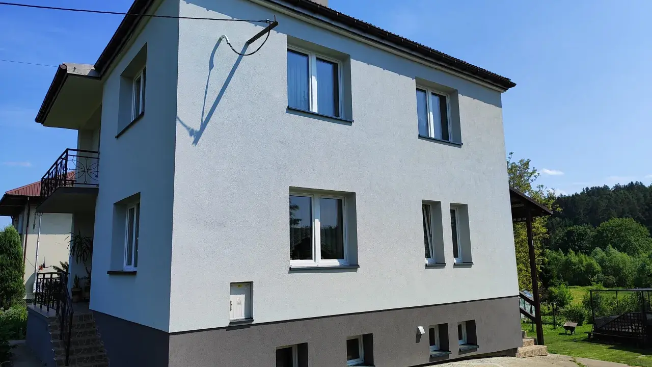 Jasny dom typu kostka z szarym cokołem i balkonem, otoczony zielenią.