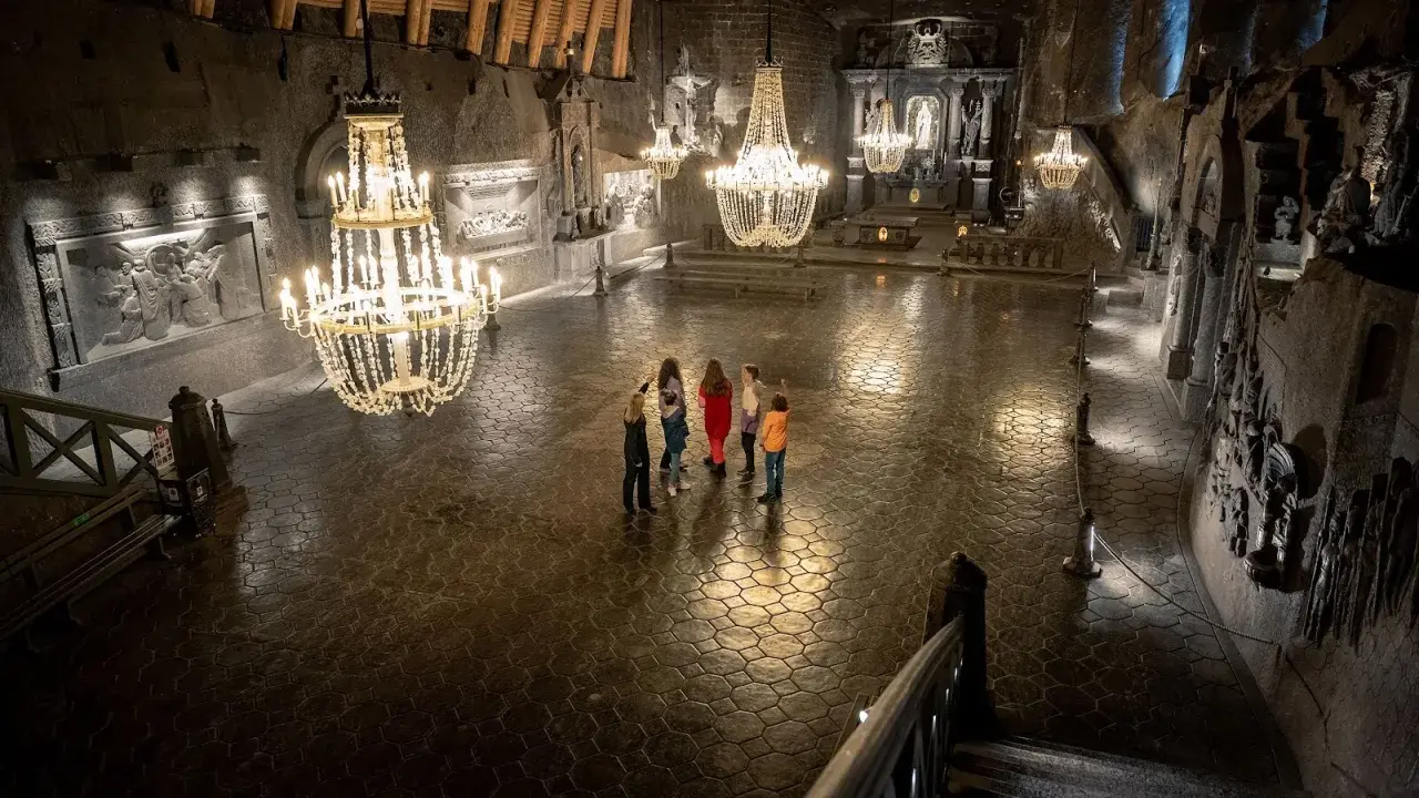 Grupa turystów w podziemnej kaplicy Kopalni Soli w Wieliczce, oświetlonej kryształowymi żyrandolami.