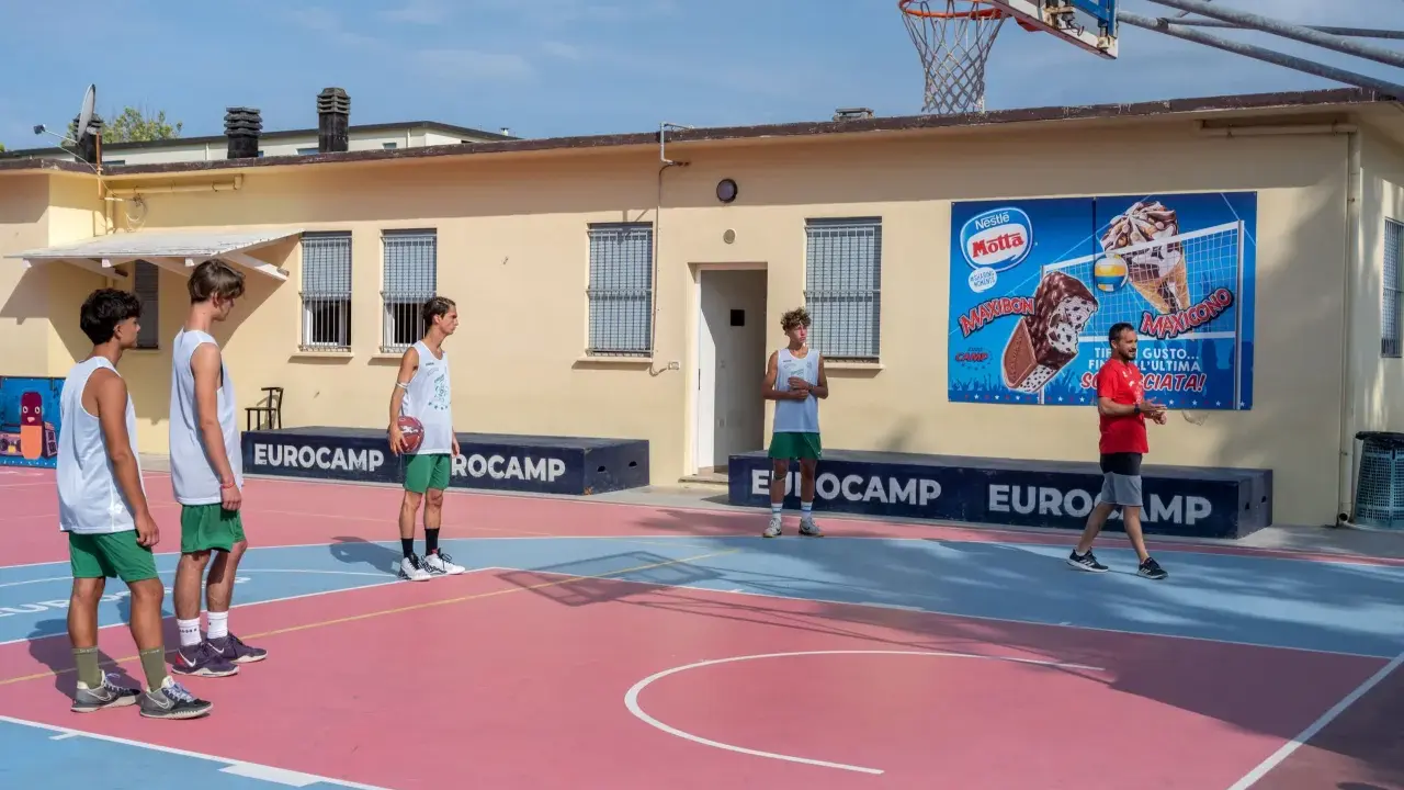 Ragazzi in tenuta sportiva giocano a basket in un camp estivo a Cesenatico, con un cartellone pubblicitario di gelati Motta sullo sfondo.