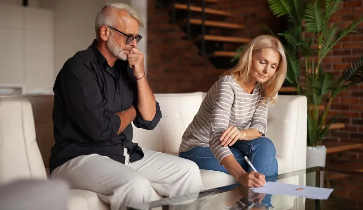 Un couple signe des documents, peut-être une désolidarisation de bail, dans un salon moderne. L'homme réfléchit, la femme écrit.
