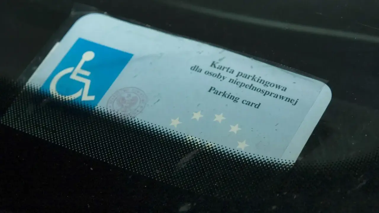 Niebieska karta parkingowa dla osoby niepełnosprawnej z symbolem wózka inwalidzkiego, umieszczona za przednią szybą samochodu.