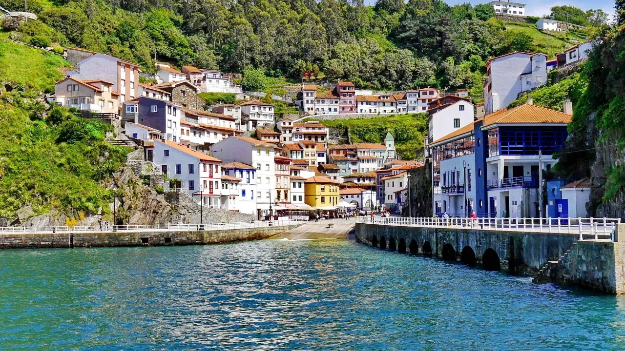 Bunte Häuser schmiegen sich an einen grünen Hang in Cudillero, Spanien. Das türkisfarbene Wasser des Hafens spiegelt die malerische Kulisse wider.