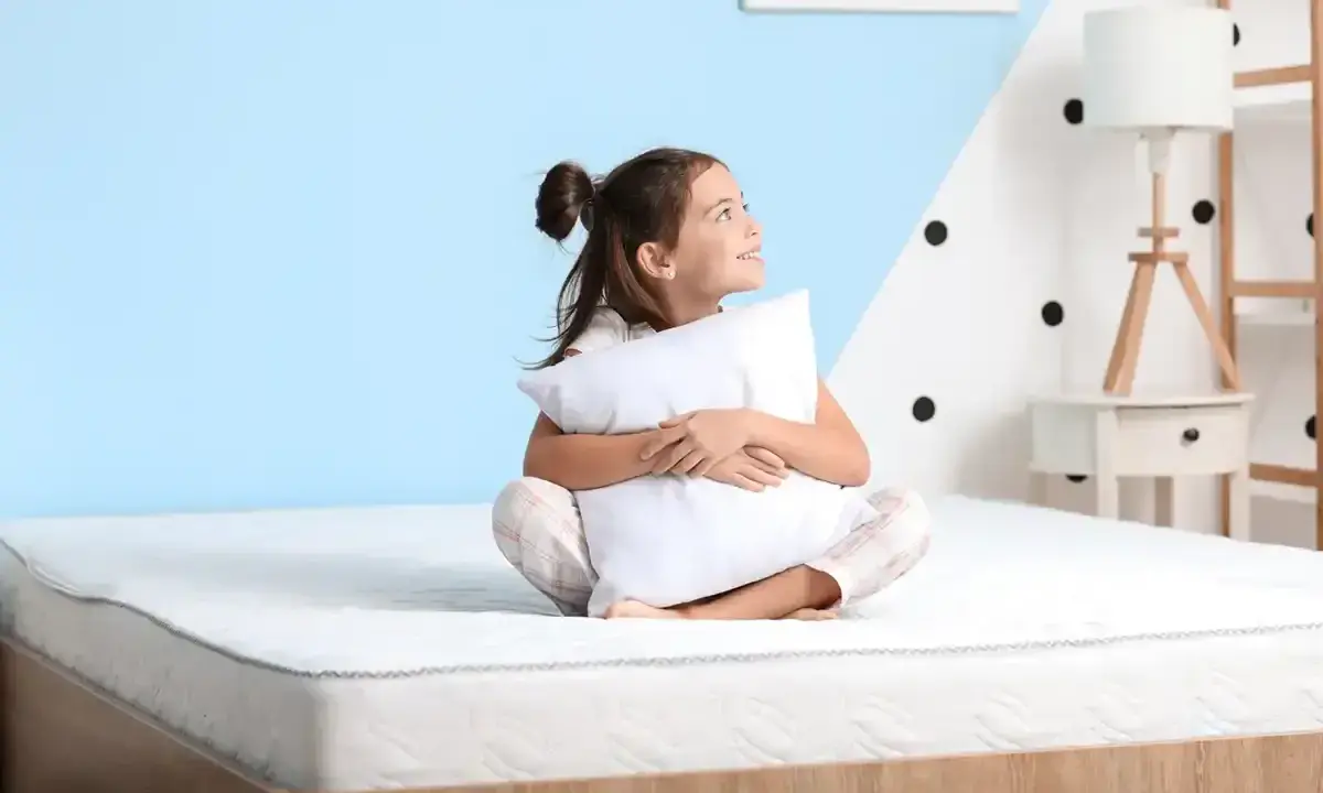 Niña sonriente abraza almohada en un colchón, pensando qué colchon es mejor para niños.