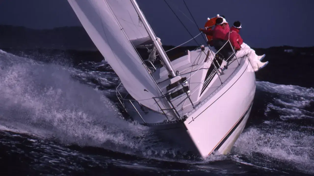 Voilier blanc fendant les vagues, deux personnes en ciré rouge. Une scène de navigation intense, digne des premiers 285 avis.