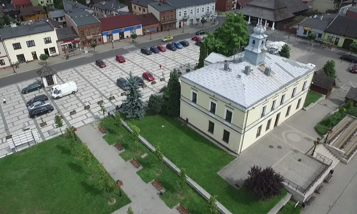 Sławków rynek z lotu ptaka: żółty budynek ratusza, brukowany plac z samochodami i zielony park z ławkami.