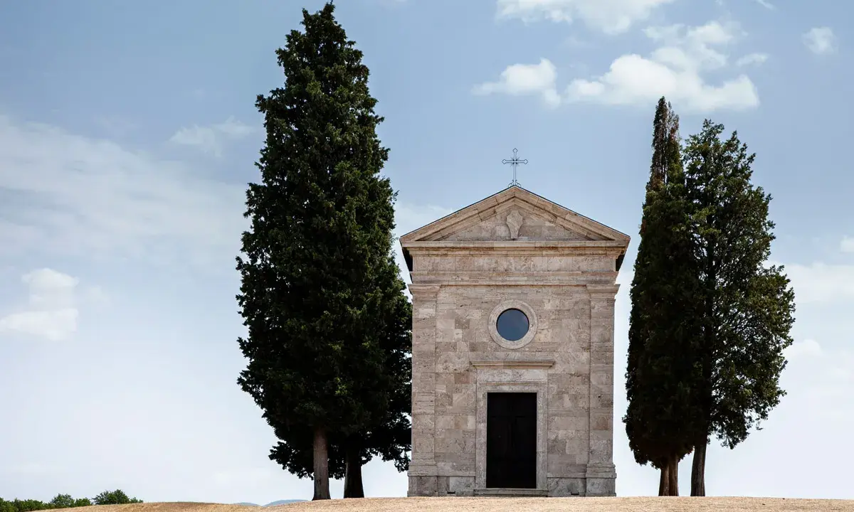 Piccola chiesa in pietra con cipressi, icona del Val d'Orcia, sotto un cielo azzurro con nuvole bianche.