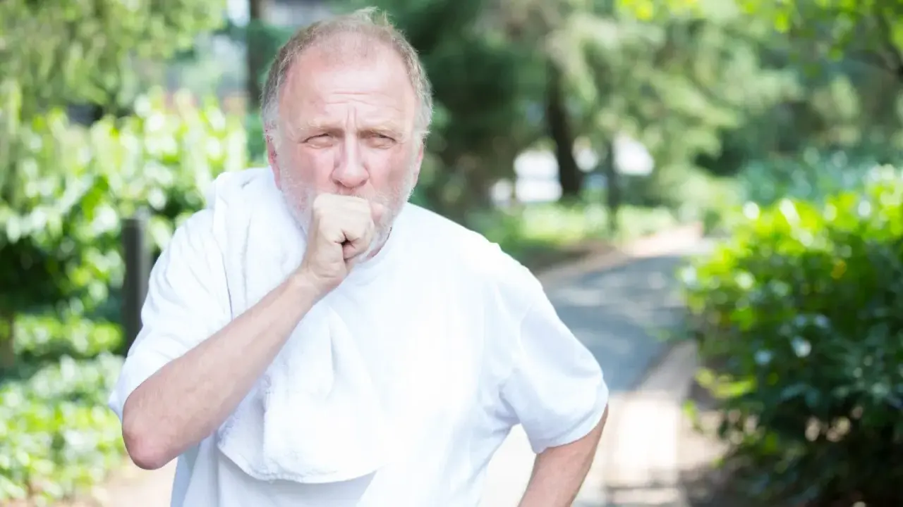 Mann mit COPD hustet im Freien. Sein dicker Bauch ist durch die weiße Kleidung sichtbar.